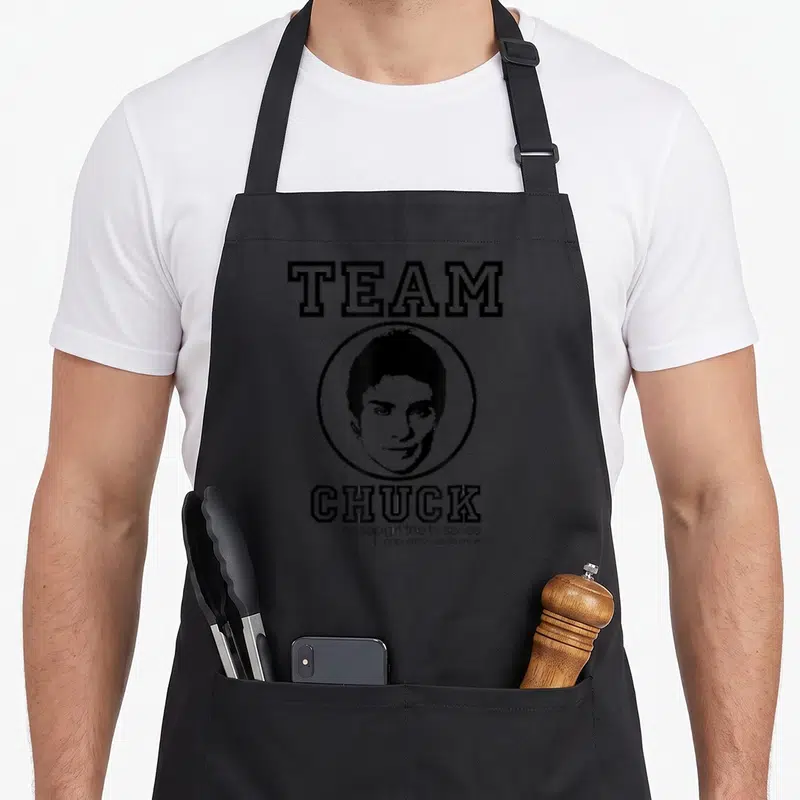 Funny chuck gossip apron - Black and white circular portrait of a man with TEAM CHUCK text and show slogan — shown on a black cotton canvas apron against a white background