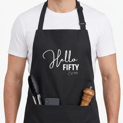 50th hello fifty est birthday apron - White Hello FIFTY Est 1973 stacked typography on grey background — shown on a black cotton canvas apron against a white background