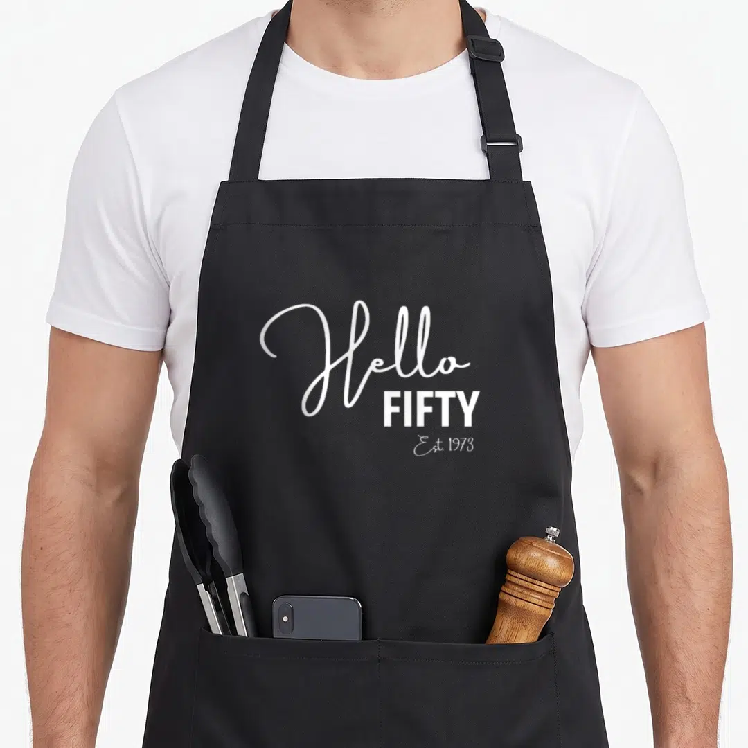 50th hello fifty est birthday apron - White Hello FIFTY Est 1973 stacked typography on grey background — shown on a black cotton canvas apron against a white background