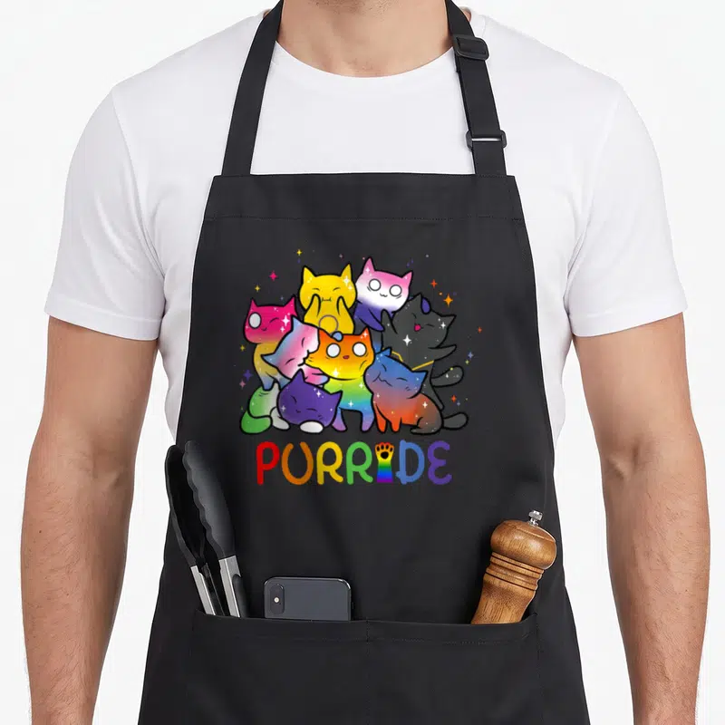 Purride animal apron - Rainbow cats clustered above PURRIDE with paw-print I, cartoon style — shown on a black cotton canvas apron against a white background