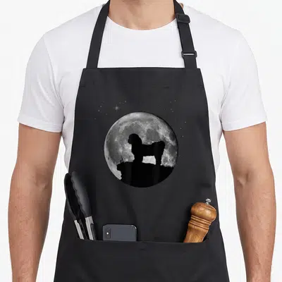 Chrysanthemum dog apron - Black dog silhouette standing on rock in front of full moon and stars — shown on a black cotton canvas apron against a white background
