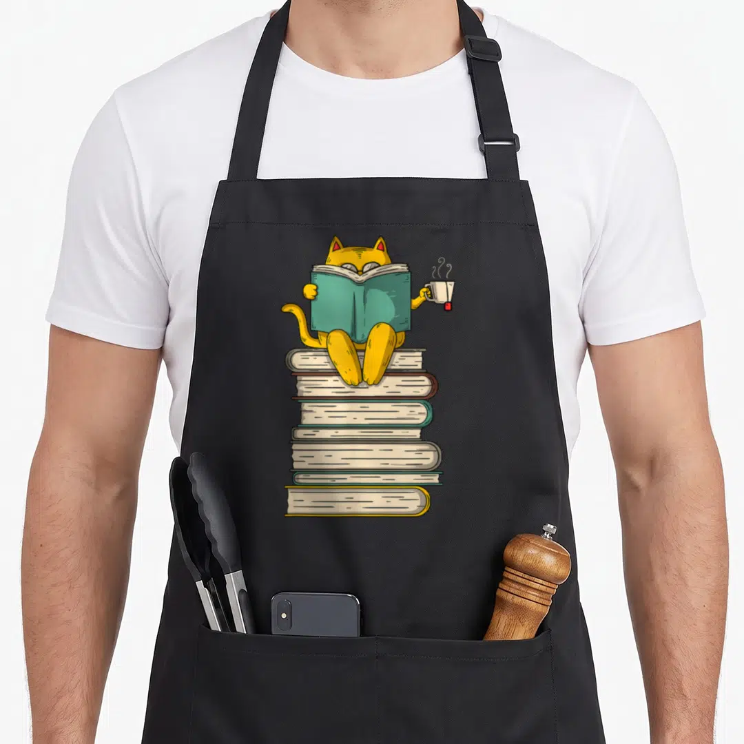 Book cat apron - yellow cartoon cat reading teal book on stacked books holding white mug — shown on a black cotton canvas apron against a white background
