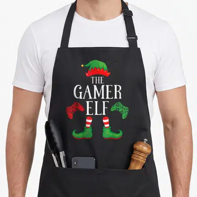 Gamer xmas christmas elf apron - Stacked text THE GAMER ELF with red and green controllers and elf hat and shoes — shown on a black cotton canvas apron against a white background