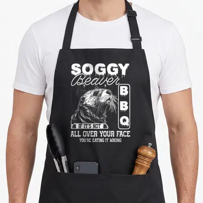 Eating beaver apron - White distressed beaver head with dripping SOGGY BBQ text and pun slogan — shown on a black cotton canvas apron against a white background