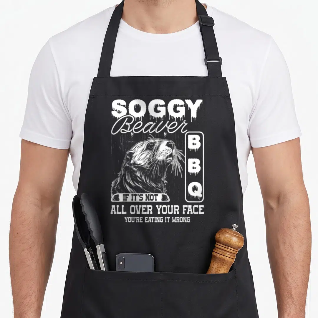 Eating beaver apron - White distressed beaver head with dripping SOGGY BBQ text and pun slogan — shown on a black cotton canvas apron against a white background