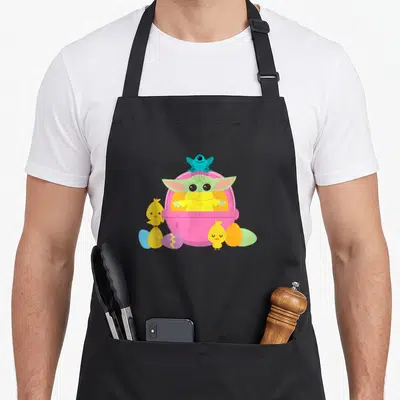 Friend easter apron - Grogu in pink pod with blue creature perched atop and yellow chicks with colorful eggs — shown on a black cotton canvas apron against a white background