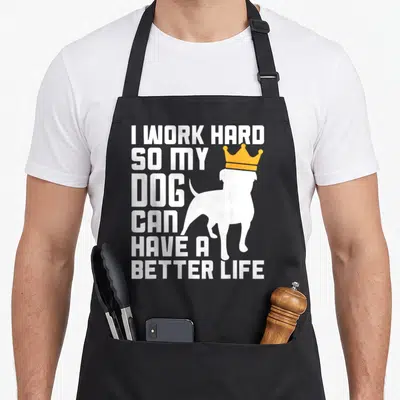 Hard pup apron - Stacked white text I work hard so my dog can have a better life next to a crowned dog silhouette — shown on a black cotton canvas apron against a white background