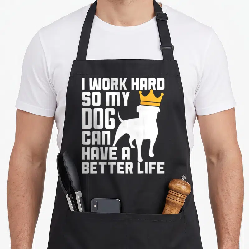 Hard pup apron - Stacked white text I work hard so my dog can have a better life next to a crowned dog silhouette — shown on a black cotton canvas apron against a white background