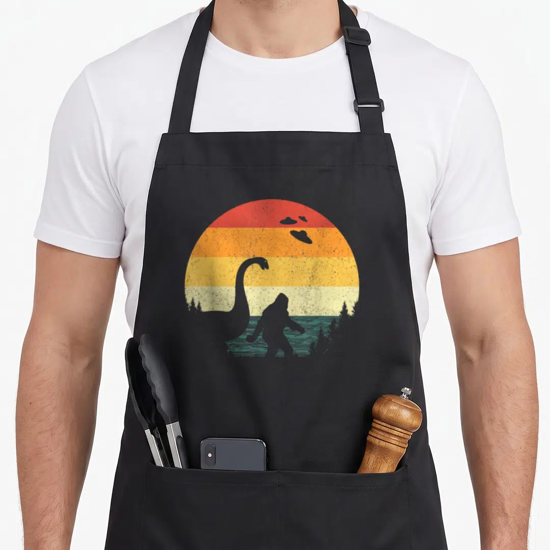 80s bigfoot apron - Grey sauropod and bigfoot silhouettes against red orange yellow and green striped semi-circle sun — shown on a black cotton canvas apron against a white background