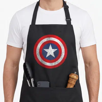 Cute shield captain apron - Distressed red white and blue circular shield with concentric rings and central white star — shown on a black cotton canvas apron against a white background