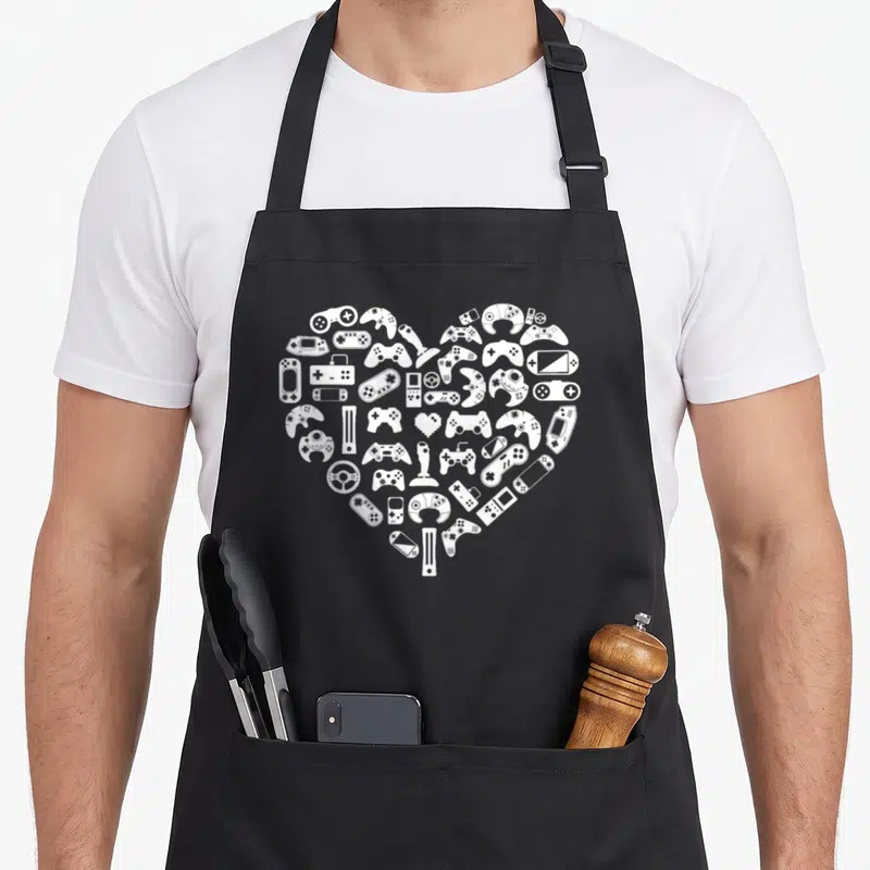 Gamer valentine's day apron - White heart shape formed by a cluster of various gaming controllers and consoles — shown on a black cotton canvas apron against a white background