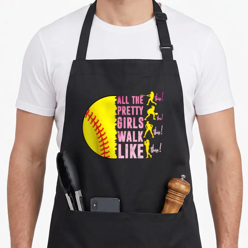 Walk pretty apron - Half yellow softball with red stitching next to pink text and four yellow player silhouettes — shown on a black cotton canvas apron against a white background