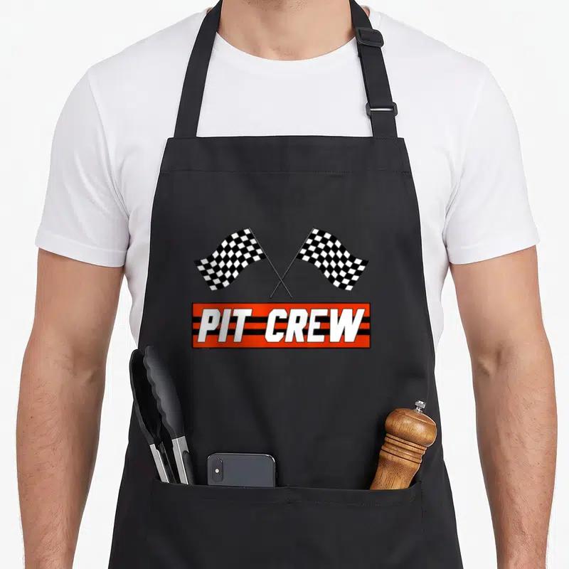 Pit car apron - Two crossed checkered flags above an orange and black striped banner reading PIT CREW in white letters — shown on a black cotton canvas apron against a white background