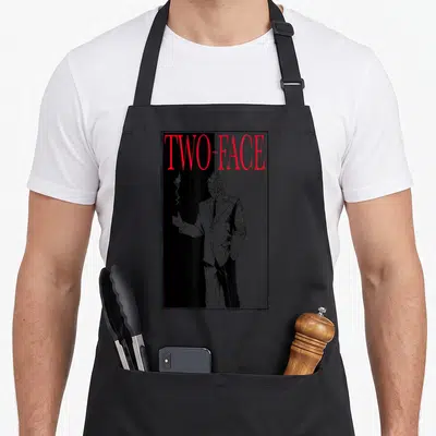 Funny hobby face apron - Two-Face character in red text and black white line art holding a coin — shown on a black cotton canvas apron against a white background