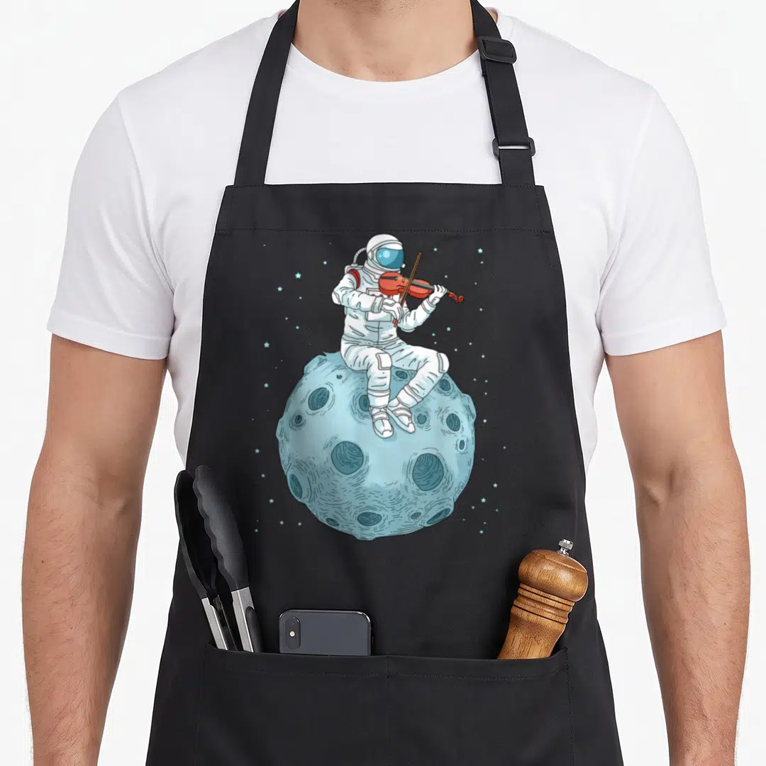 Violin fiddle apron - Cartoon astronaut in white suit playing red violin on light blue cratered moon with stars — shown on a black cotton canvas apron against a white background