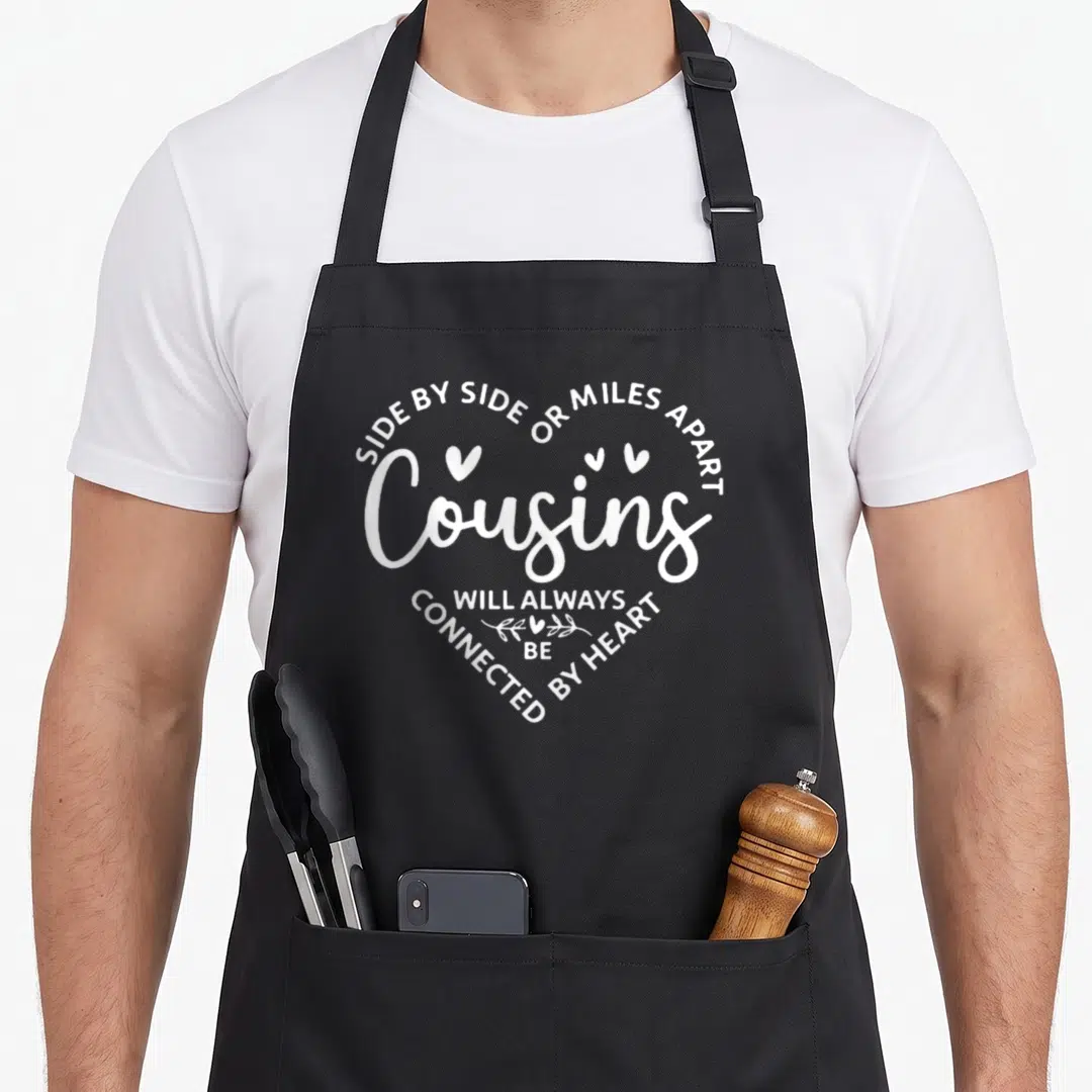 Mile apart apron or apron - White text arranged in a heart shape reading Cousins connected by heart — shown on a black cotton canvas apron against a white background