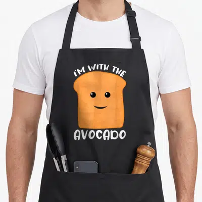 Funny avocado toast apron - Orange cartoon toast with smile and white I'M WITH THE AVOCADO text — shown on a black cotton canvas apron against a white background