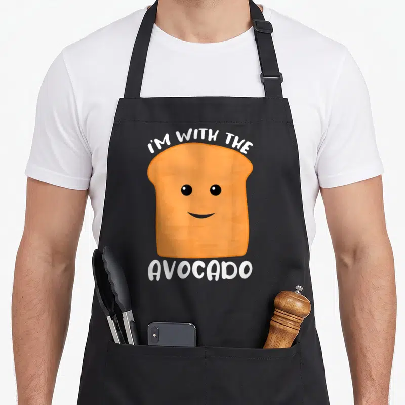 Funny avocado toast apron - Orange cartoon toast with smile and white I'M WITH THE AVOCADO text — shown on a black cotton canvas apron against a white background