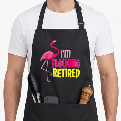 Retired flamingo apron - Pink cartoon flamingo stands beside stacked text reading I'm Flocking Retired in bold colors — shown on a black cotton canvas apron against a white background