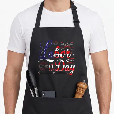 Funny labor union aprons - Labor Day text in blue white and red flag patterns with gears wrenches stars and arrow — shown on a black cotton canvas apron against a white background