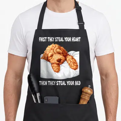 Sleeping goldendoodle apron - Ginger dog sleeping in white bedding with text about stealing hearts and beds — shown on a black cotton canvas apron against a white background
