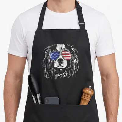 Cavalier king apron - Distressed white dog head wearing sunglasses with blue stars and red white stripes — shown on a black cotton canvas apron against a white background