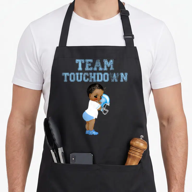 Touchdown tutu apron - Cartoon baby in blue shorts hugging a football helmet under TEAM TOUCHDOWN text — shown on a black cotton canvas apron against a white background