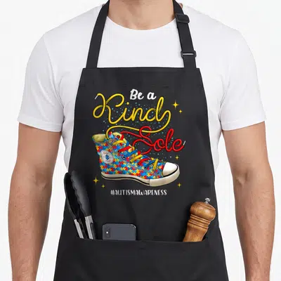 Autistic shoe apron - Sneaker with puzzle pattern and 'Be a Kind Sole' text in yellow and red — shown on a black cotton canvas apron against a white background