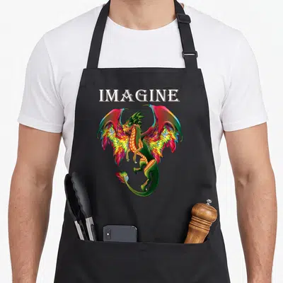 Breathing dragon apron - Colorful cartoon dragon with rainbow wings below white IMAGINE text — shown on a black cotton canvas apron against a white background
