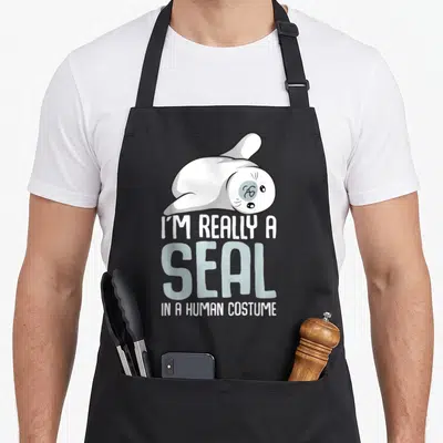 Seal lion apron - Cartoon seal lying down above stacked text I'M REALLY A SEAL IN A HUMAN COSTUME — shown on a black cotton canvas apron against a white background