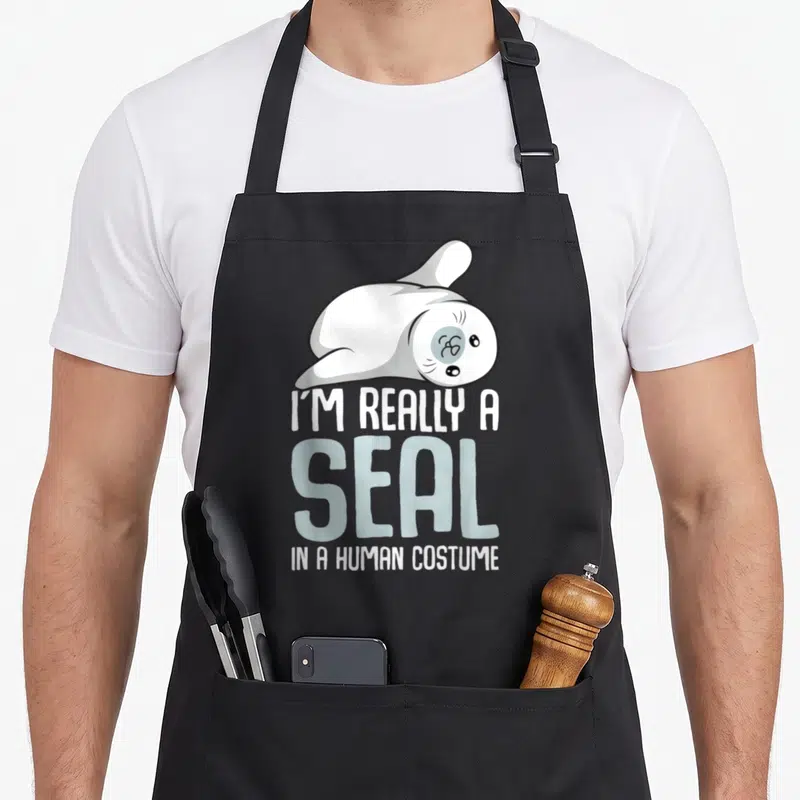 Seal lion apron - Cartoon seal lying down above stacked text I'M REALLY A SEAL IN A HUMAN COSTUME — shown on a black cotton canvas apron against a white background