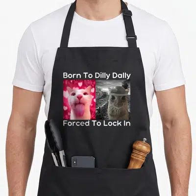 Dilly cat apron - Pink tongue-out cat next to grey helmeted cat with white meme text above and below — shown on a black cotton canvas apron against a white background