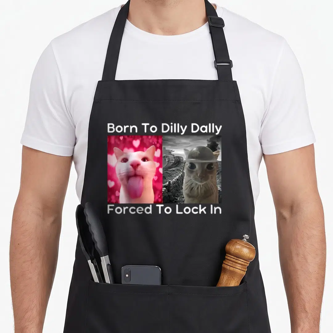 Dilly cat apron - Pink tongue-out cat next to grey helmeted cat with white meme text above and below — shown on a black cotton canvas apron against a white background