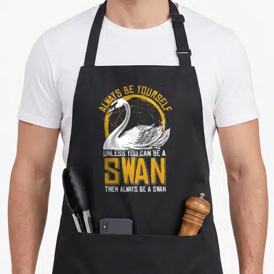 Cute animal swan apron - White swan illustration with yellow circle and text ALWAYS BE YOURSELF UNLESS YOU CAN BE — shown on a black cotton canvas apron against a white background