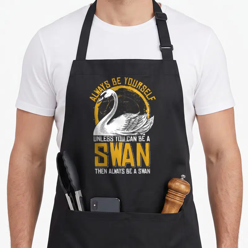 Cute animal swan apron - White swan illustration with yellow circle and text ALWAYS BE YOURSELF UNLESS YOU CAN BE — shown on a black cotton canvas apron against a white background