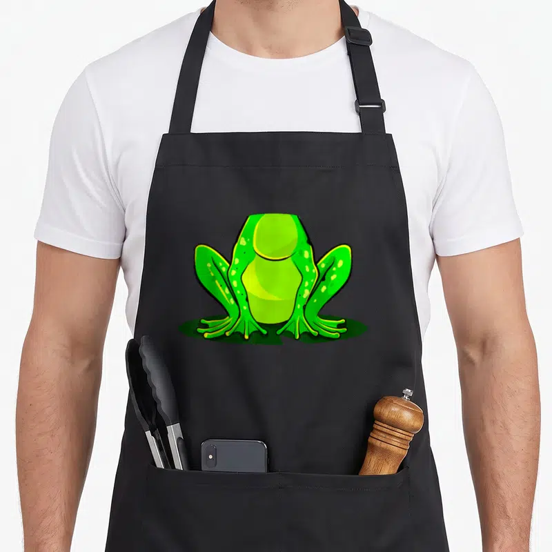 Frog halloween apron - Green cartoon frog sitting on dark green oval shadow with yellow accents — shown on a black cotton canvas apron against a white background