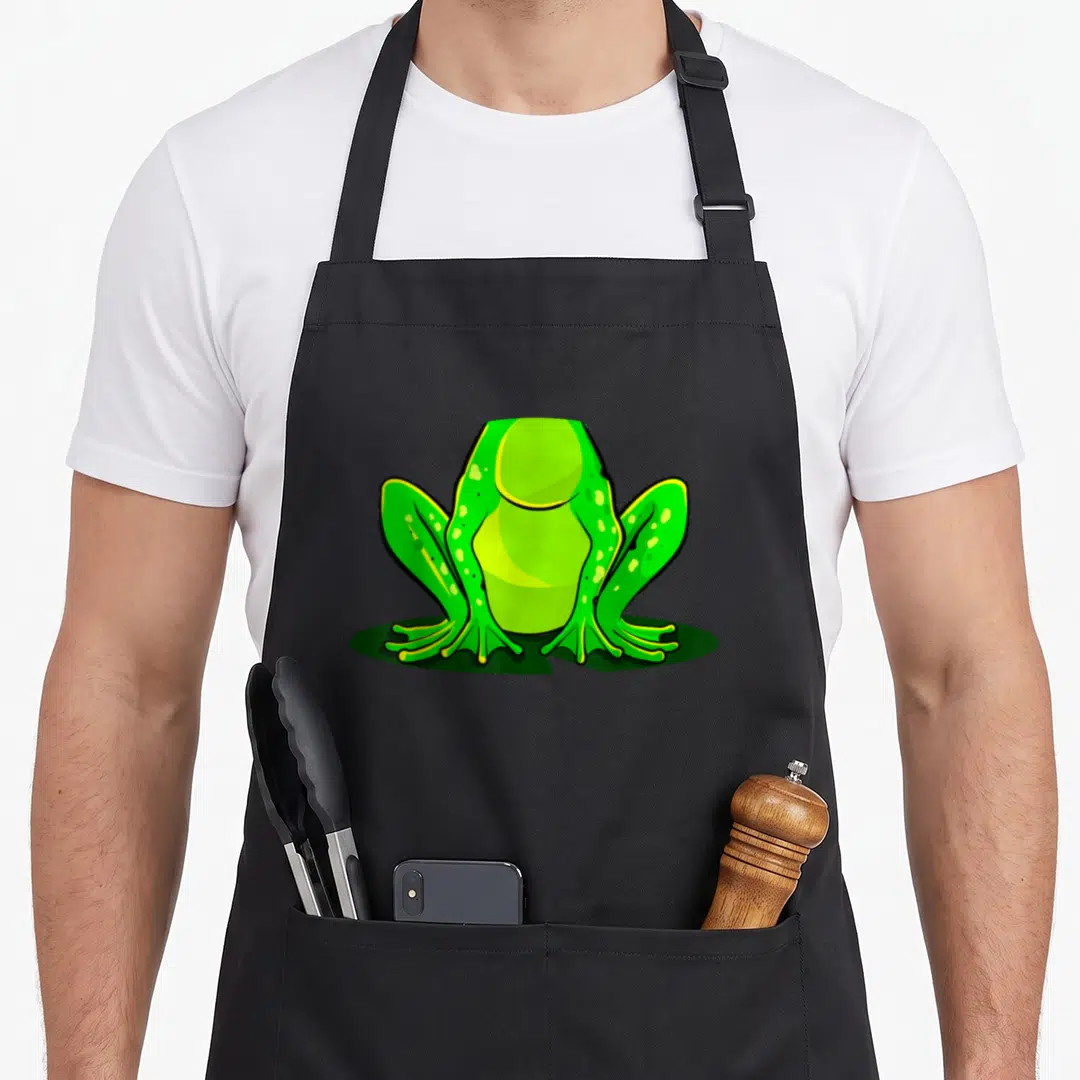 Frog halloween apron - Green cartoon frog sitting on dark green oval shadow with yellow accents — shown on a black cotton canvas apron against a white background
