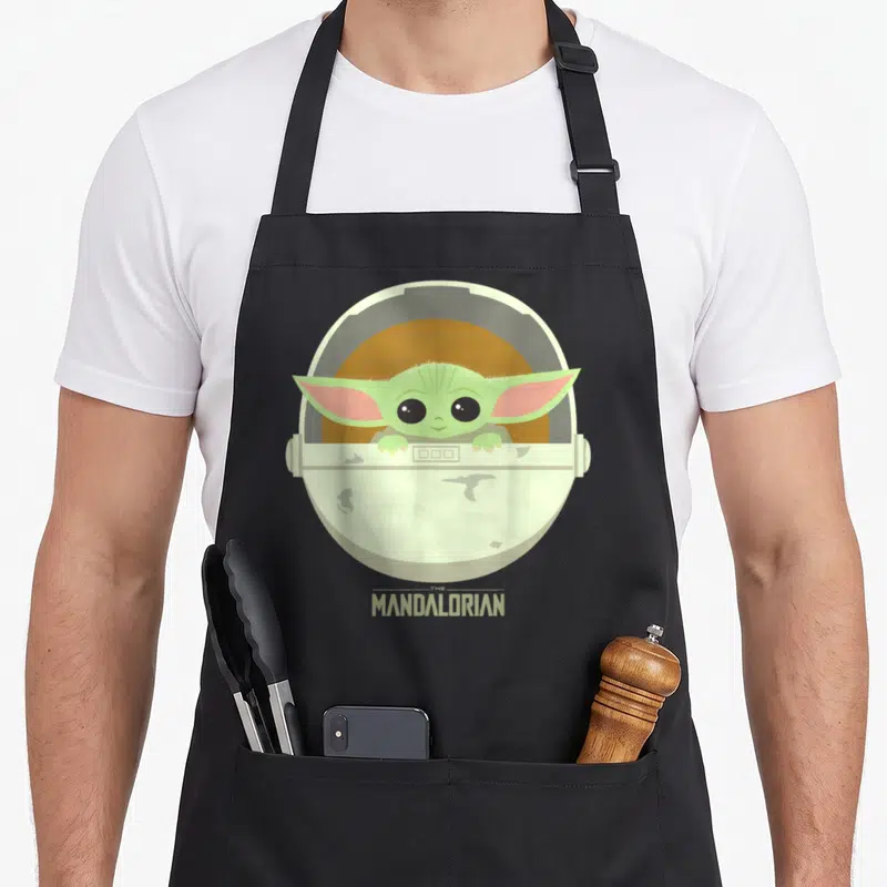 Portrait bassinet apron - Grogu in circular pod with THE MANDALORIAN text below on brown background — shown on a black cotton canvas apron against a white background