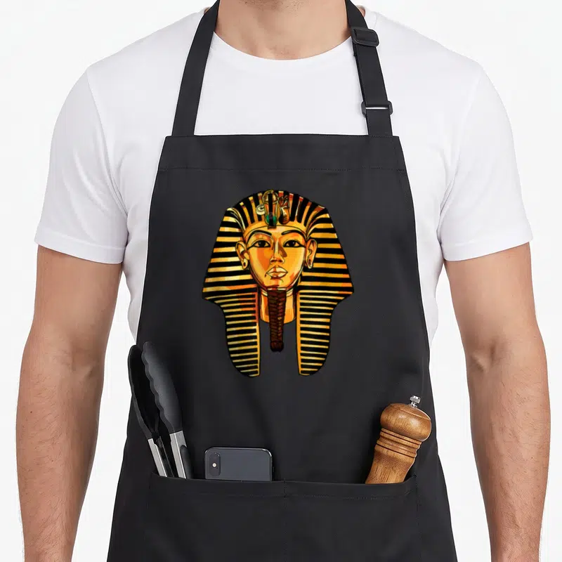 King egyptian apron - Cartoon pharaoh head with striped nemes headcloth and false beard in gold, black, and orange — shown on a black cotton canvas apron against a white background