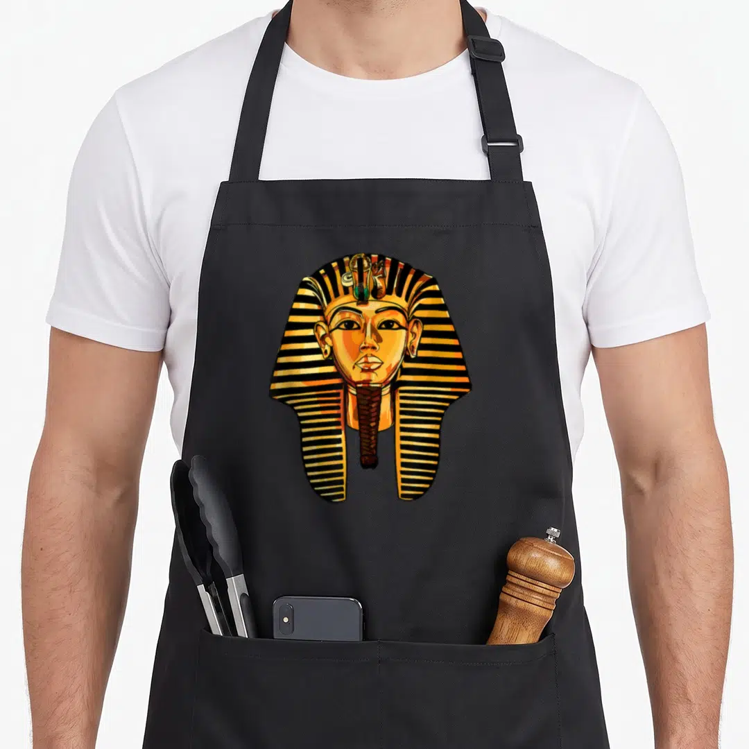 King egyptian apron - Cartoon pharaoh head with striped nemes headcloth and false beard in gold, black, and orange — shown on a black cotton canvas apron against a white background