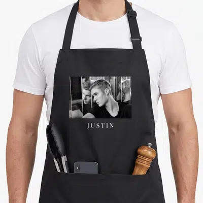 Justin observation apron - Grayscale photo of Justin Bieber with vertical reflections and name JUSTIN below — shown on a black cotton canvas apron against a white background