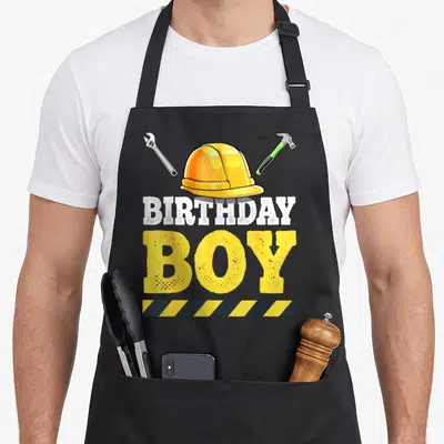 Funny birthday construction apron - Yellow hard hat, wrench, hammer, BIRTHDAY BOY text, hazard stripe — shown on a black cotton canvas apron against a white background