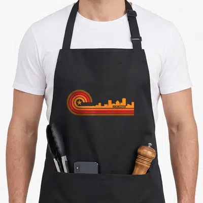 Cityscape rochester apron - Retro Rochester skyline with brown red orange yellow stripes and star on left — shown on a black cotton canvas apron against a white background