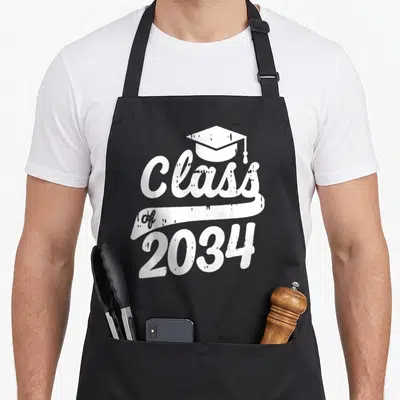 Class kindergarten apron - White graduation cap above Class of 2034 text on a distressed ribbon banner — shown on a black cotton canvas apron against a white background