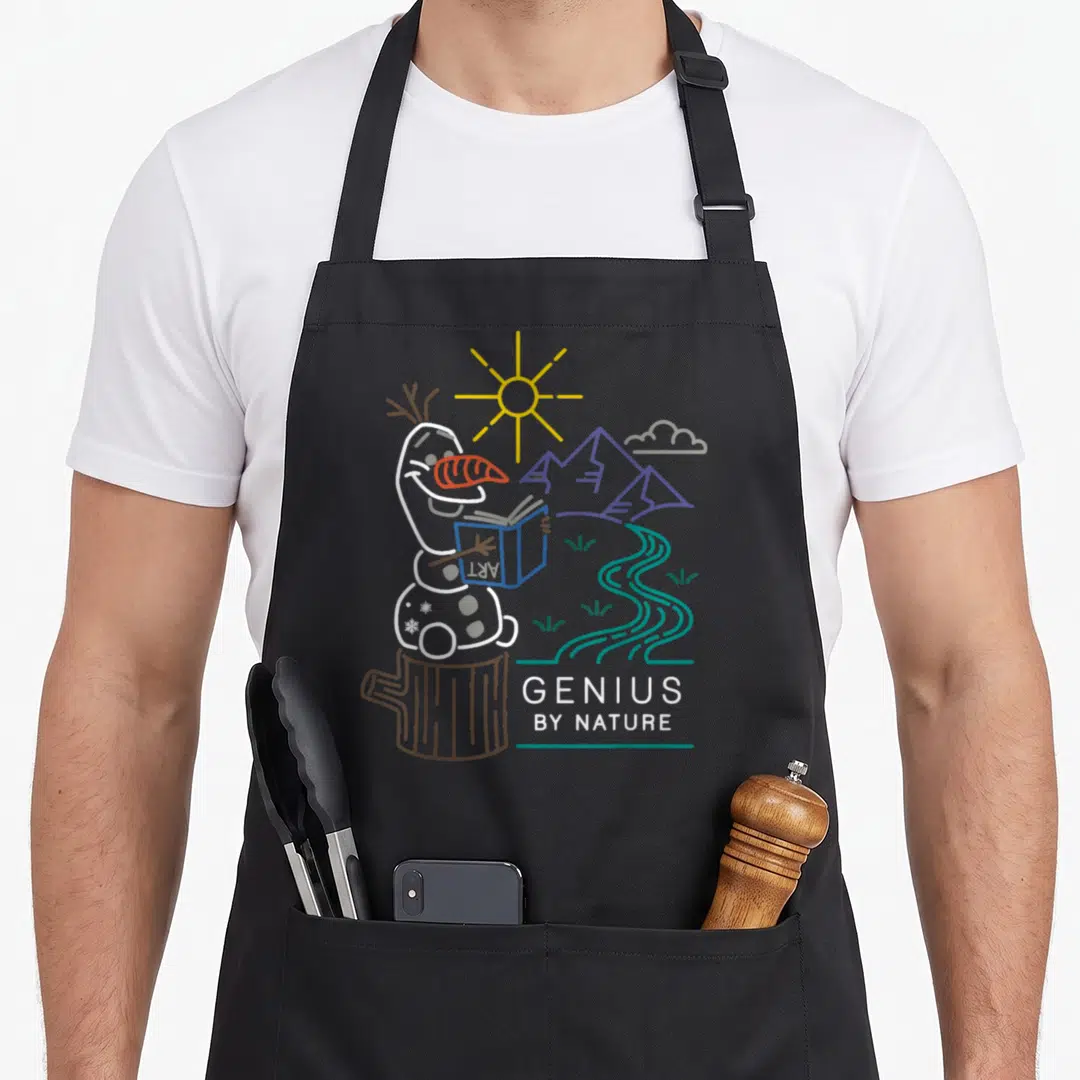 Genius frozen apron - Snowman reading book on stump with GENIUS BY NATURE text and landscape — shown on a black cotton canvas apron against a white background