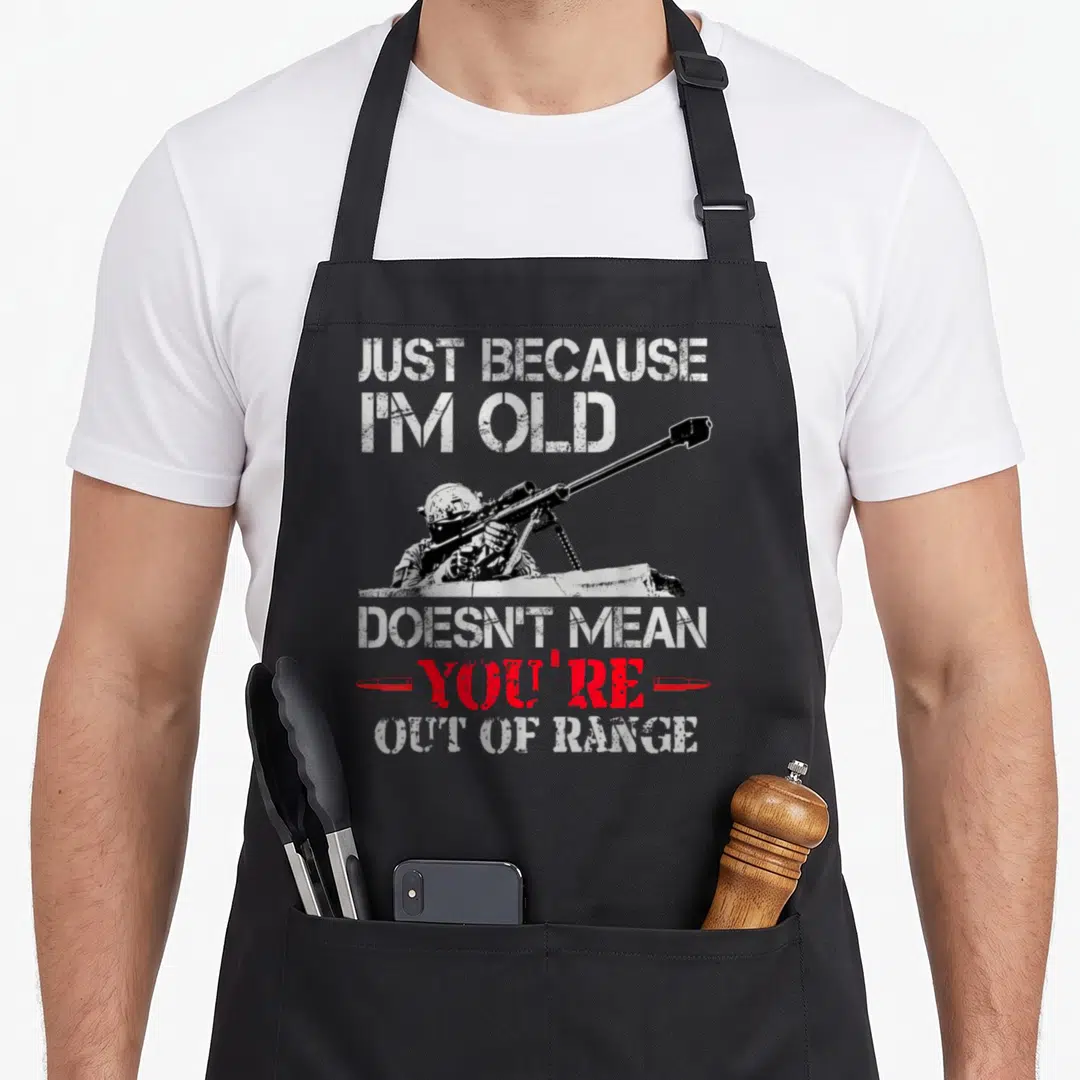 Mean the range apron - Distressed white text reading JUST BECAUSE I'M OLD above a black and white sniper holding — shown on a black cotton canvas apron against a white background