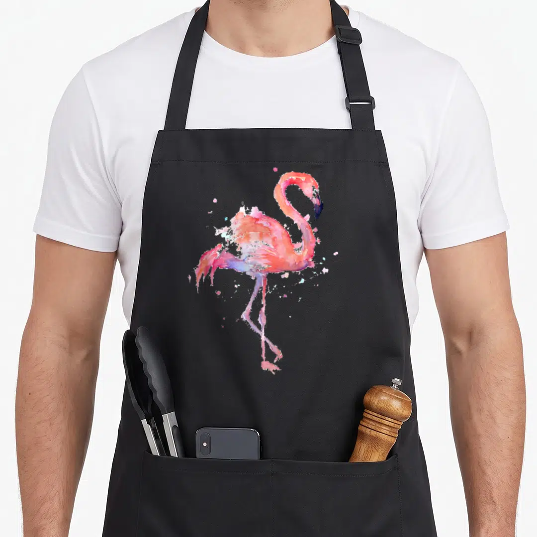Bird pink flamingo apron - Watercolor flamingo in pink, orange, and purple with paint splatters on black background — shown on a black cotton canvas apron against a white background