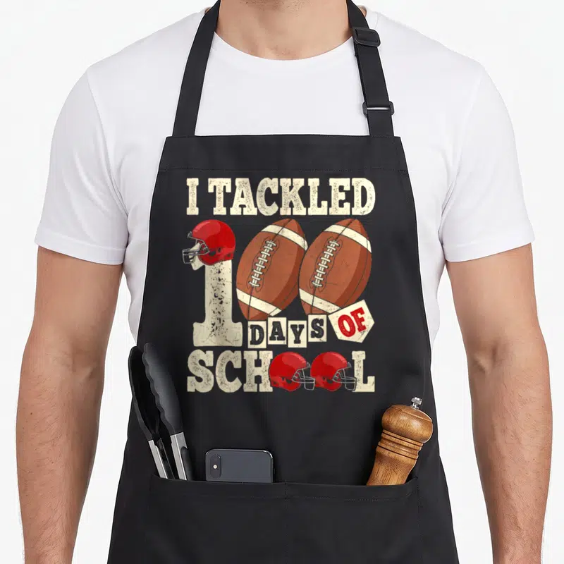 100th football apron - I Tackled 100 Days of School text with footballs and red helmets in distressed cream and brown — shown on a black cotton canvas apron against a white background