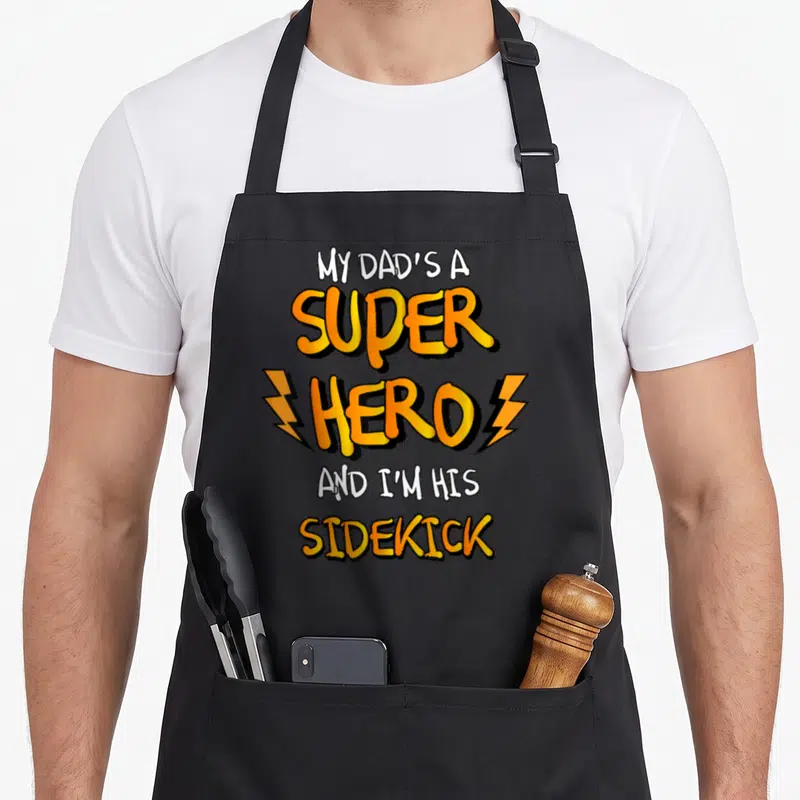 Funny superhero apron - Cartoon text MY DAD'S A SUPER HERO AND I'M HIS SIDEKICK with orange lightning bolts — shown on a black cotton canvas apron against a white background