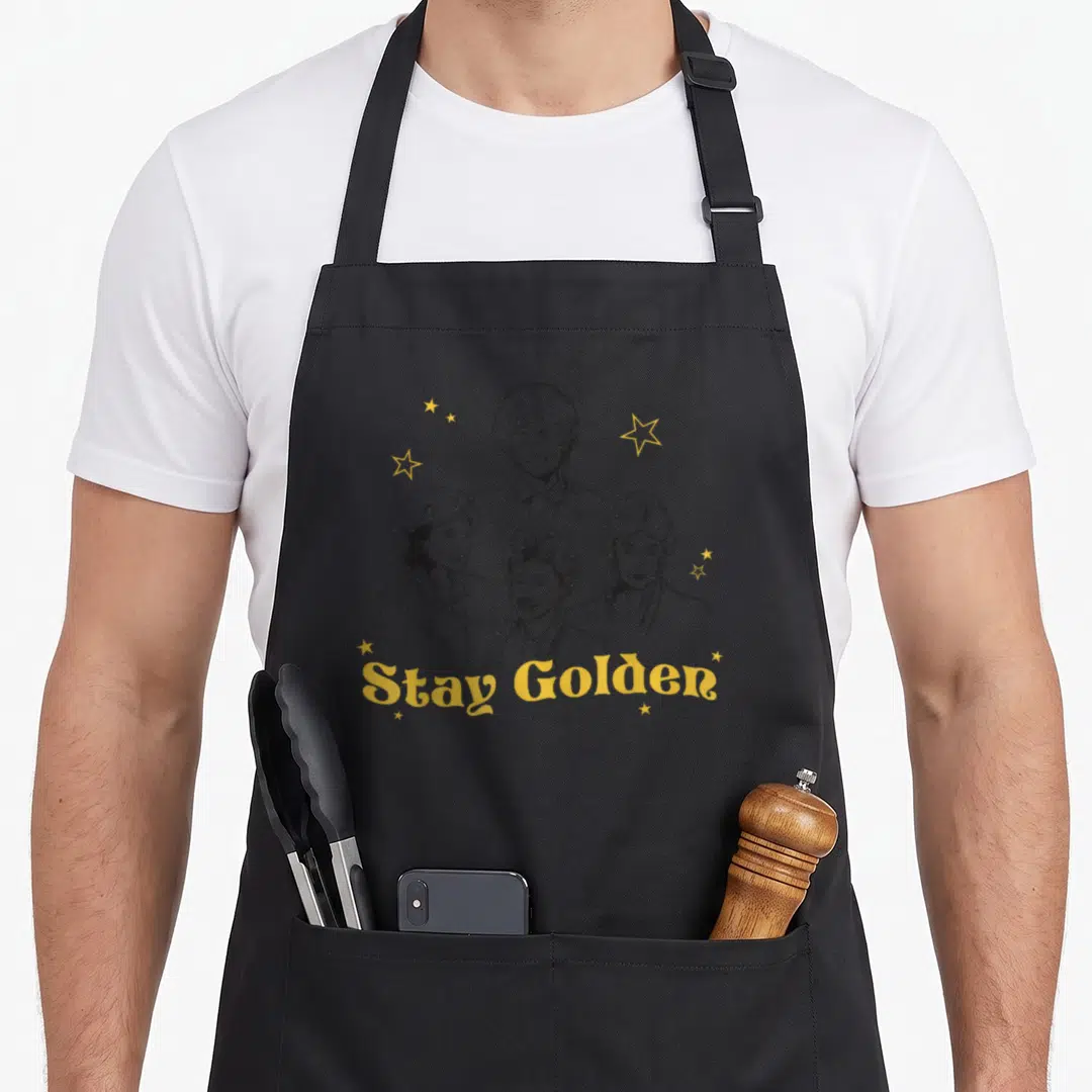 Cool stay golden apron - Four women line art portraits with yellow stars and Stay Golden text below — shown on a black cotton canvas apron against a white background